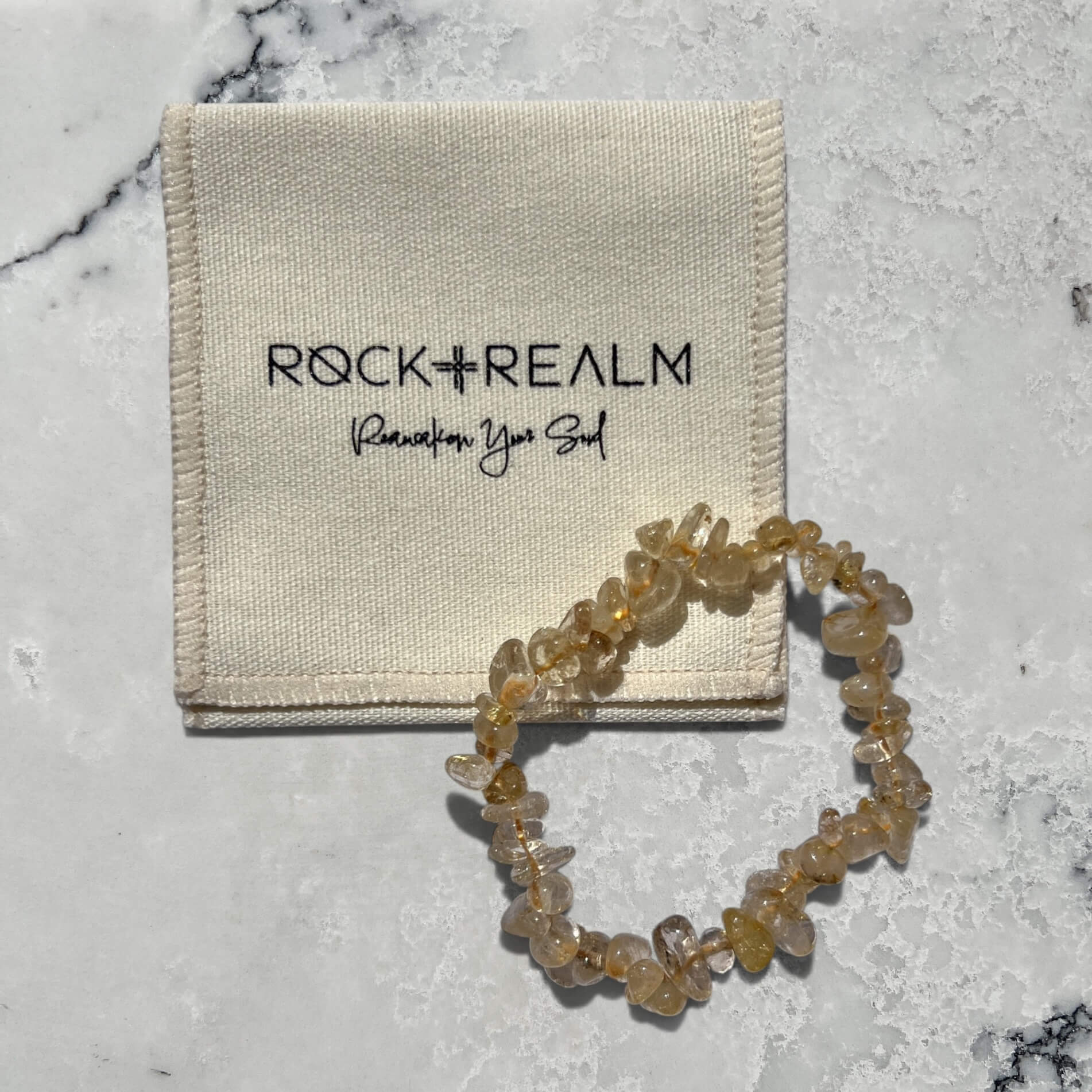 Yellow and clear golden rutilated quartz crystal chip bracelet on a marble surface with cotton 'Rock + Realm' pouch