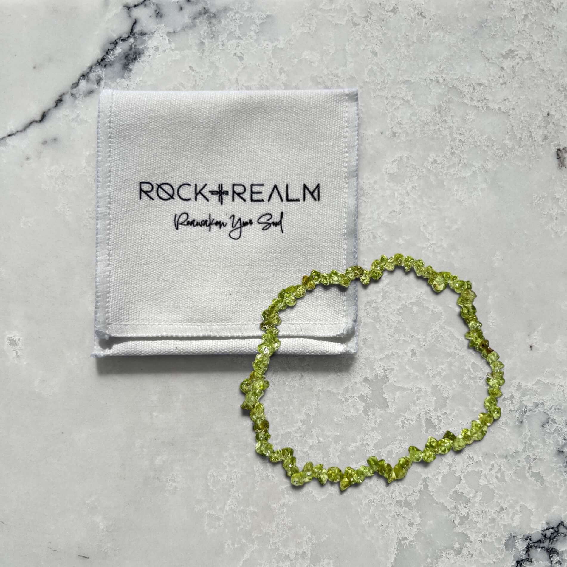 A bright green peridot crystal chip bracelet on a marble surface with a 'Rock + Realm' branded white cotton pouch