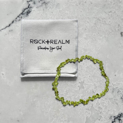 A bright green peridot crystal chip bracelet on a marble surface with a 'Rock + Realm' branded white cotton pouch