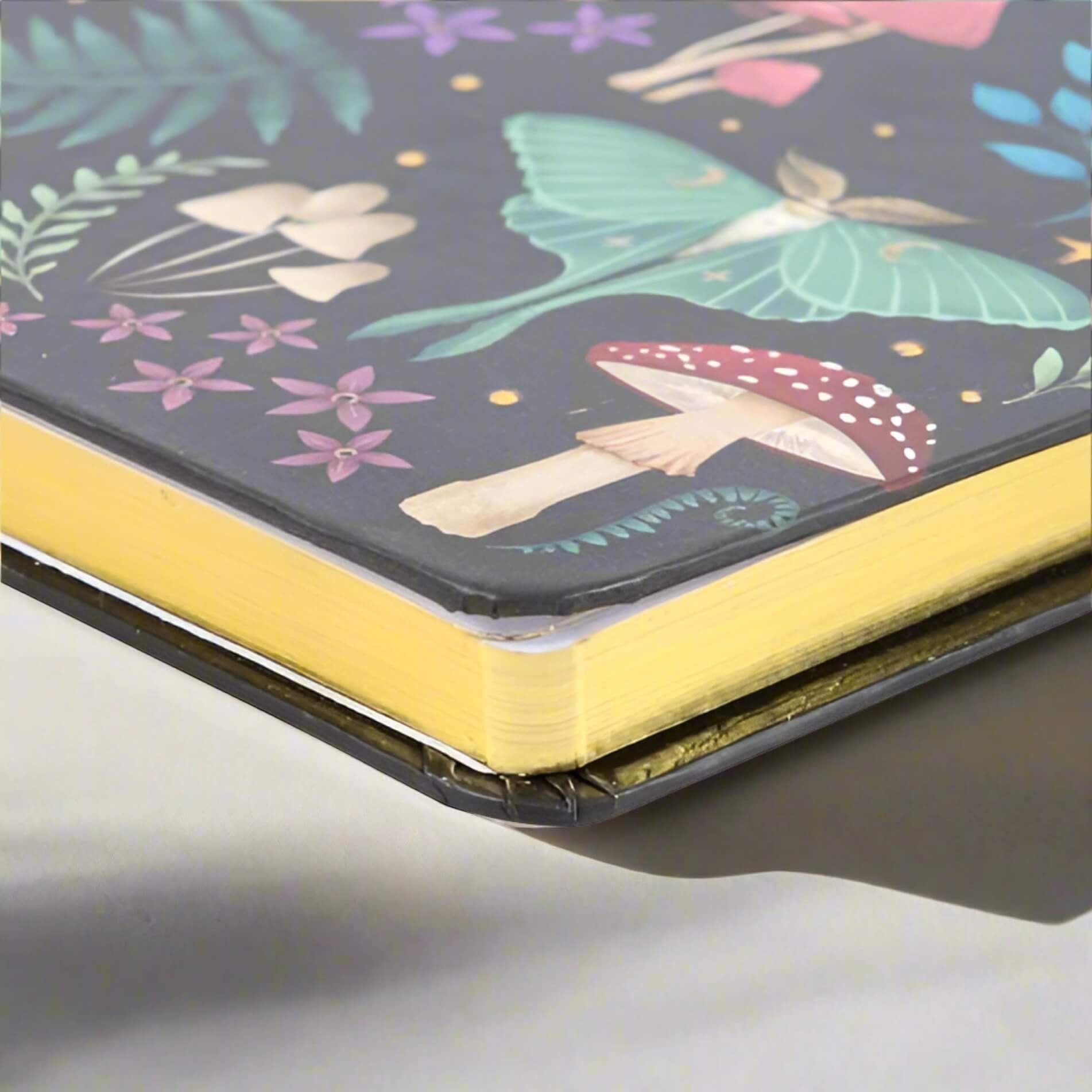 Rock + Realm Crystals Dark Forest A5 journal with a luna moth, mushrooms, and floral illustrations on a dark background. Features gold-gilded page edges.