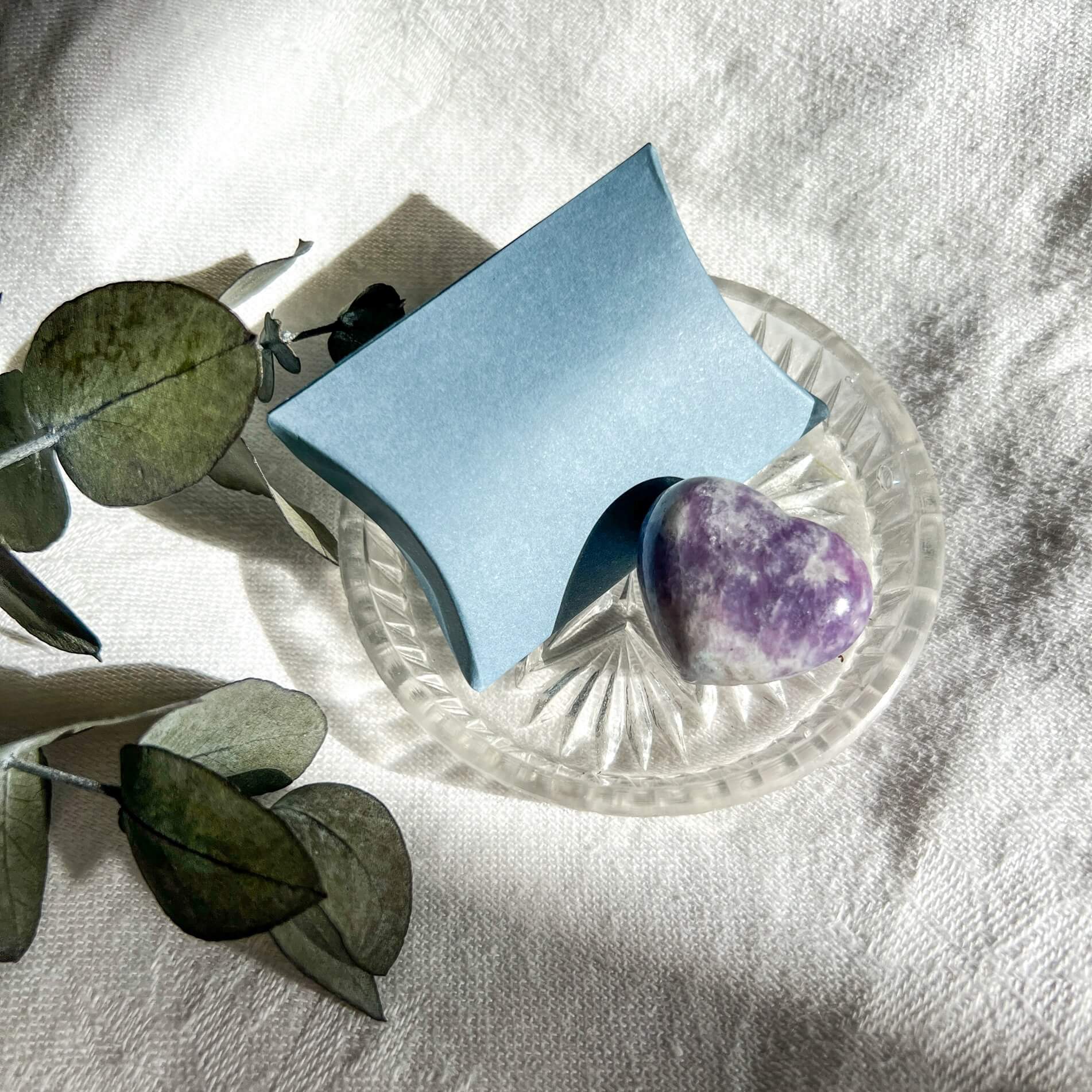 A Purple and lilac coloured lepidolite crystal mini heart displayed in a small glass dish with a pale blue pillow box and eucalyptus leaves in the background