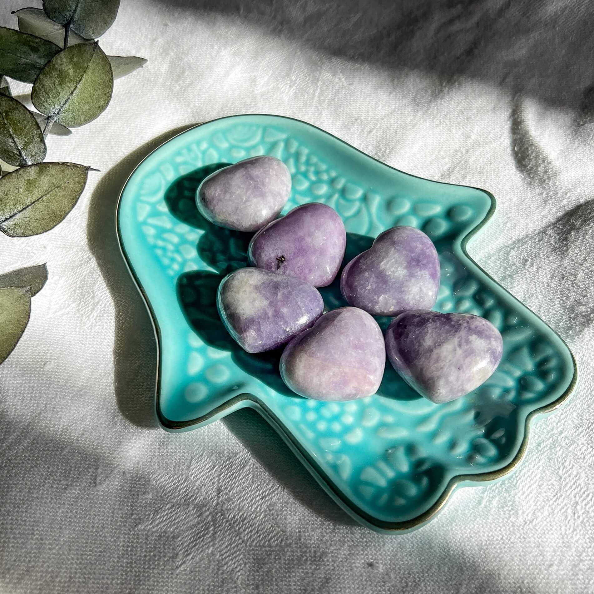 Six purple and lilac coloured lepidolite crystal mini hearts displayed in a turquoise hamsa hand trinket dish
