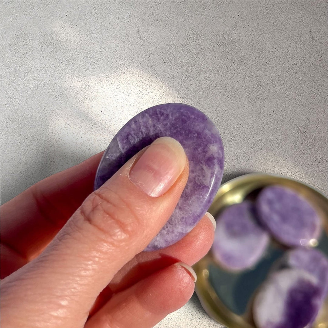 Rock + Realm Crystals Lepidolite worry stone with a thumb indentation, smooth purple with white marbling and mica inclusions pinched between thumb and finger