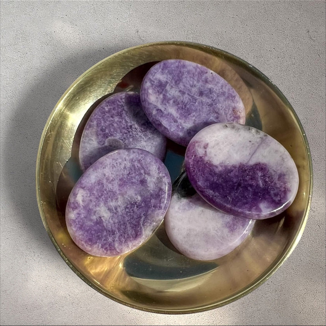 Rock + Realm Crystals Lepidolite worry stone, smooth purple stone with white marbling and mica inclusions, oval shape with thumb indentation shown from above in a brass dish