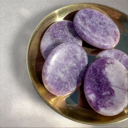Rock + Realm Crystals Lepidolite worry stone, smooth purple with white marbling and mica inclusions, oval shape with thumb indentation in a brass dish