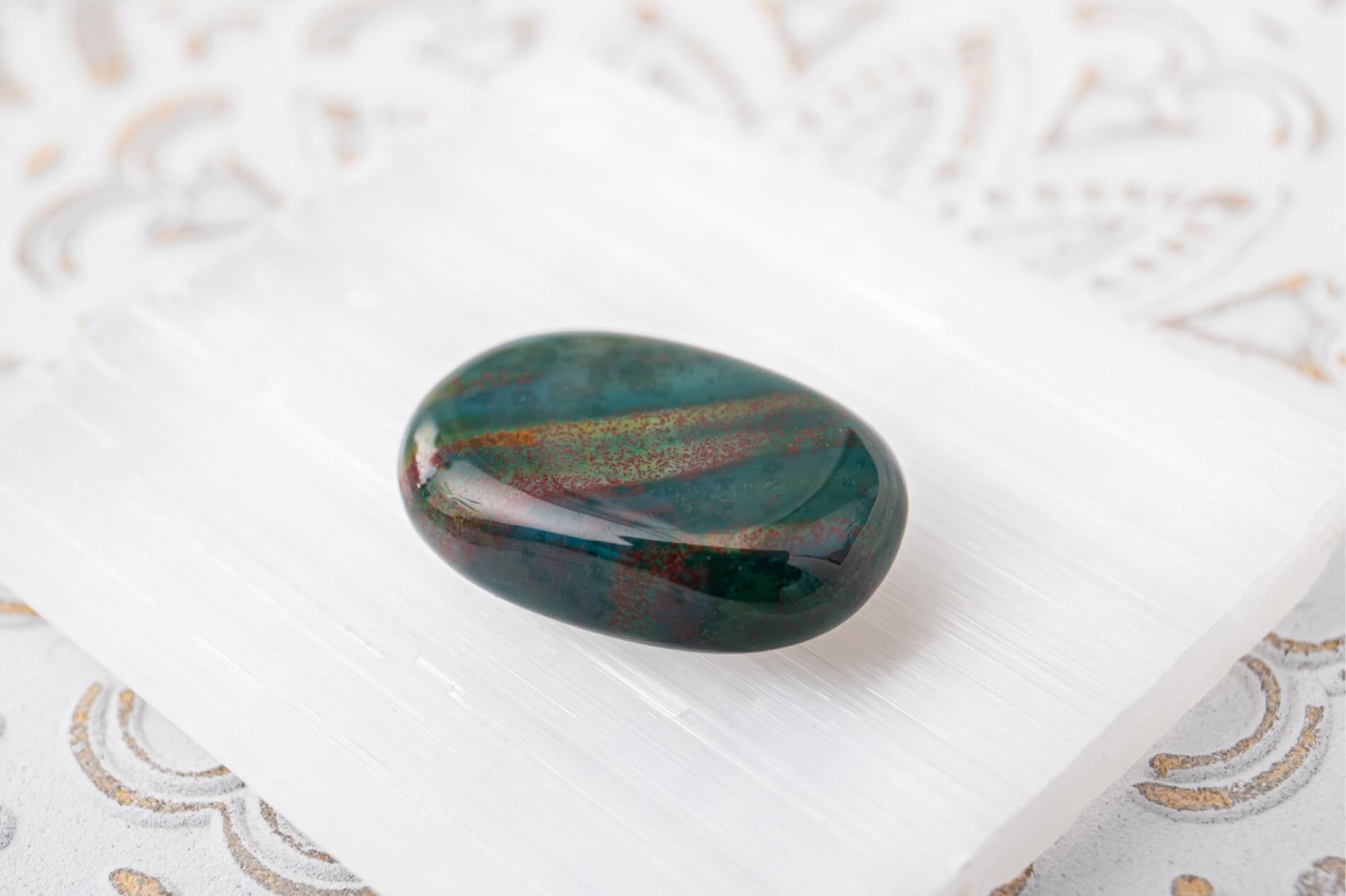 A polished dark green ova-shaped bloodstone crystal palm stone with red and yellow speckles against a dark green, resting on a white selenite slab over a white patterned fabric background.