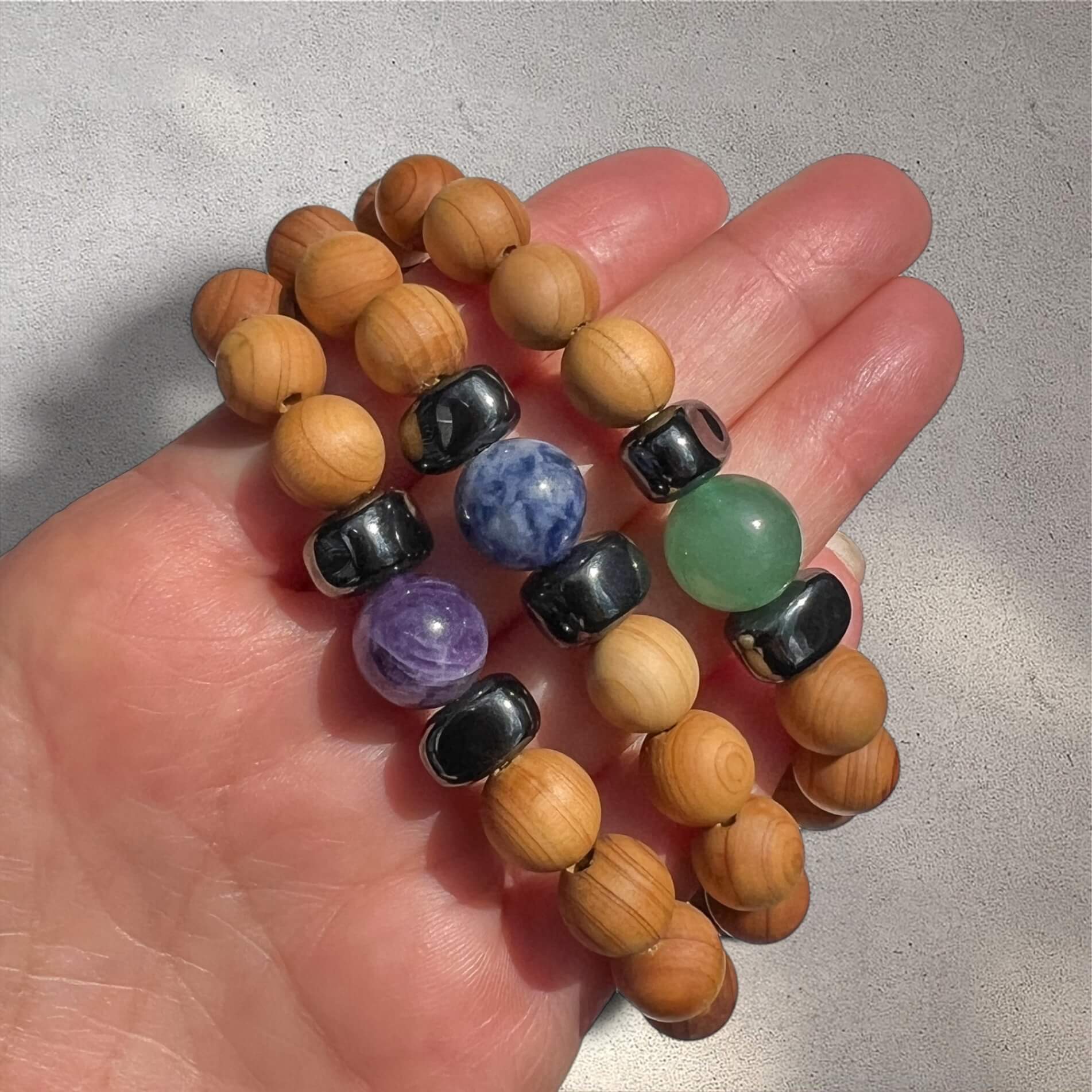 Hand holding a stack of Cedar wood beaded bracelets each with a central crystal bead in either purple amethyst, green aventurine or blue sodalite against a neutral textured background