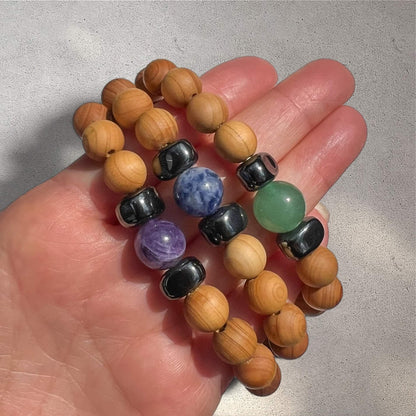 Hand holding a stack of Cedar wood beaded bracelets each with a central crystal bead in either purple amethyst, green aventurine or blue sodalite against a neutral textured background