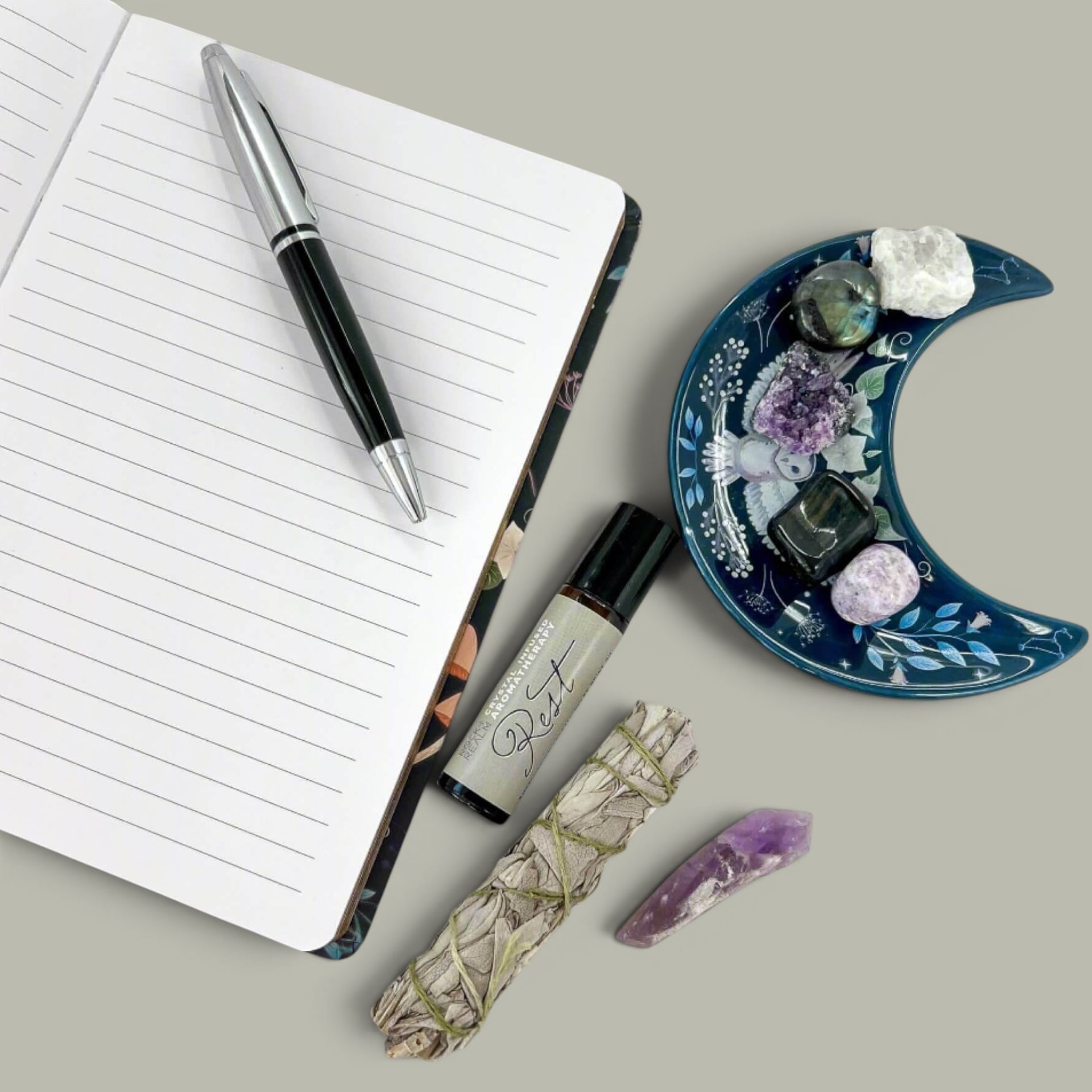 Rock + Realm mystical owl journal with lined pages, a silver and black pen, aromatherapy roller, sage bundle, and crystals on a moon-shaped dish.