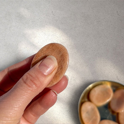 Rock + Realm Crystals peach moonstone worry stone, oval polished crystal with thumb indentation for comfort and stress relief held between thumb and forefinger
