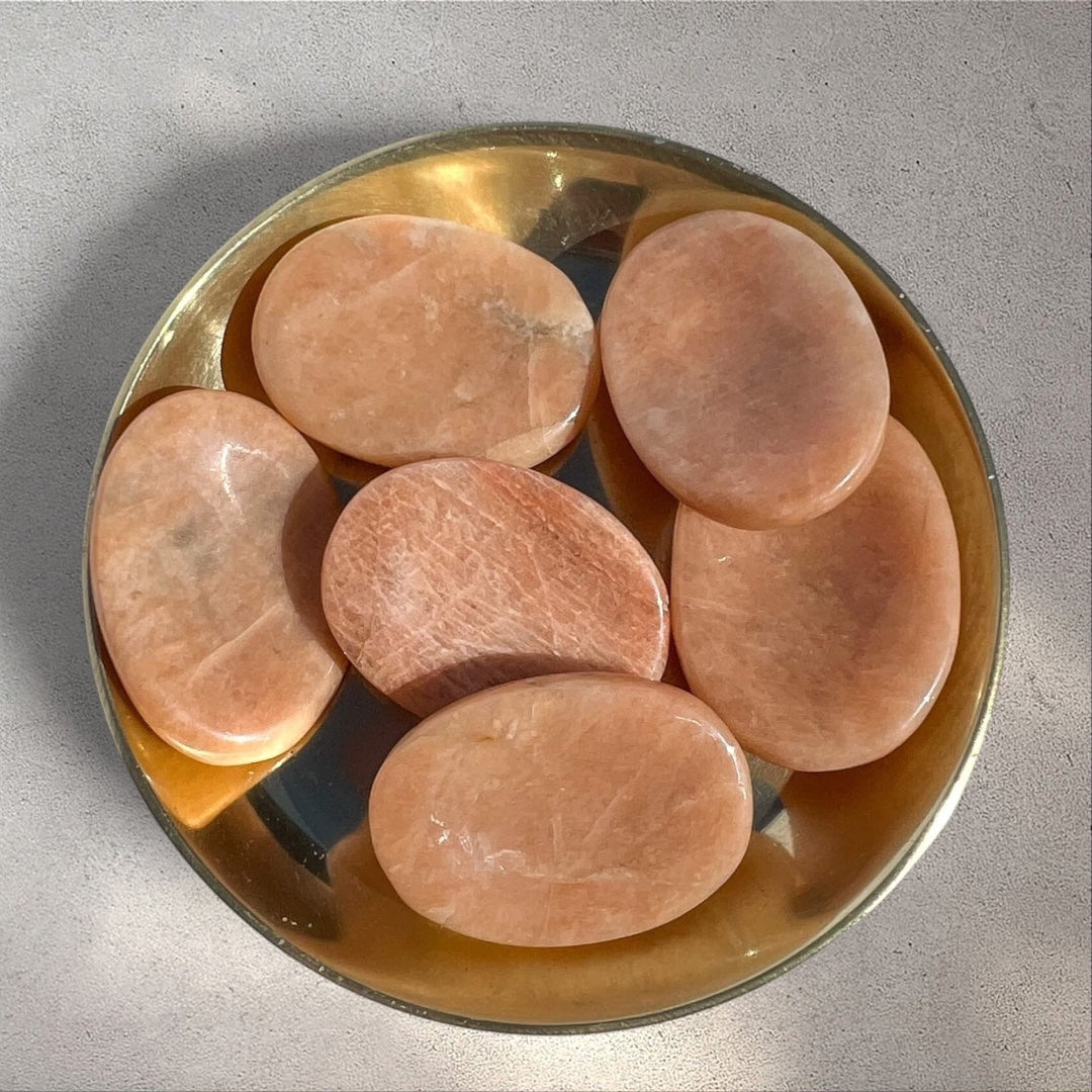 Rock + Realm Crystals polished oval peach moonstone worry stones with thumb indentation, displayed in a brass dish.