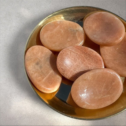 Rock + Realm Crystals polished oval peach moonstone worry stone with thumb indentation for stress relief and emotional support in a brass dish
