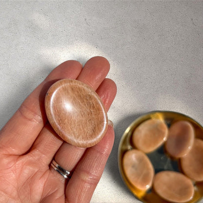 Rock + Realm Crystals polished oval peach moonstone worry stone with thumb indentation, held in hand.