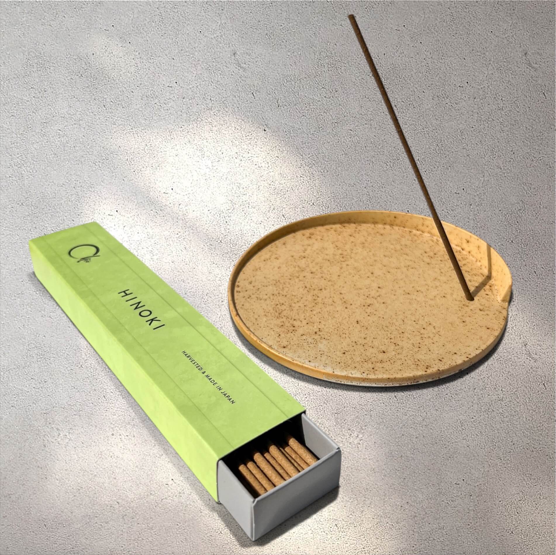 A box of hinoki incense sticks shown next to a round ceramic incense holder with one incense stick inserted, on a textured grey concrete surface.