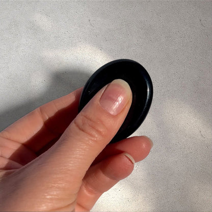 Rock + Realm Crystals black obsidian worry stone with thumb indentation, held between thumb and forefinger.