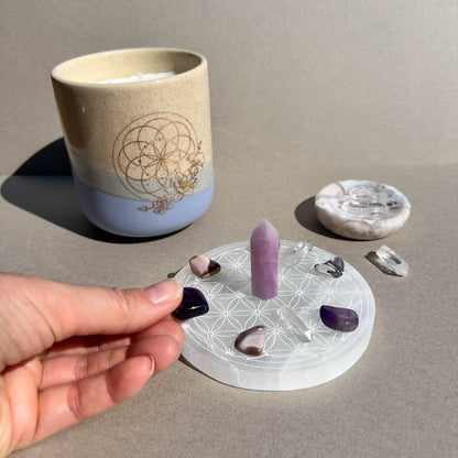 A hand holds a purple amethyst crystal placing it in a crystal grid on a white selenite plate with a sacred geometry pattern with a kunzite crystal point in its centre and other polished crystals, beside a ceramic candle and a small flower agate dish on a neutral surface.