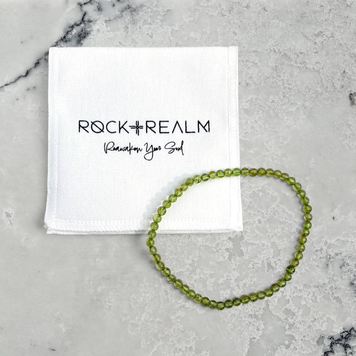 A green Peridot beaded bracelet arranged in a circle next to a white branded cotton pouch that reads ROCK + REALM Reawaken Your Soul, placed on a light marble surface.