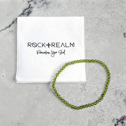 A green Peridot beaded bracelet arranged in a circle next to a white branded cotton pouch that reads ROCK + REALM Reawaken Your Soul, placed on a light marble surface.
