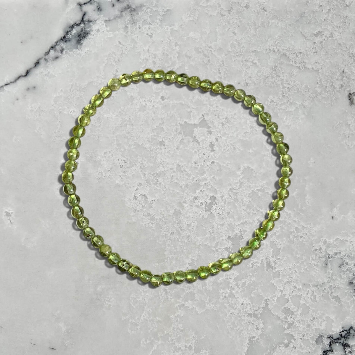 A beaded stretch bracelet made of round light green Peridot gemstone beads, arranged in a circle on a white marble surface.