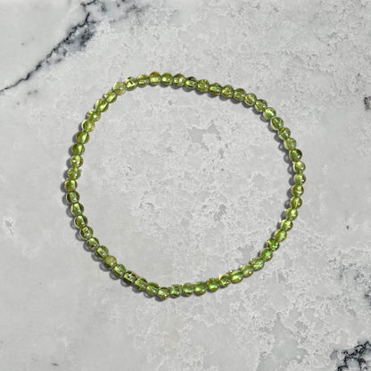 A beaded stretch bracelet made of round light green Peridot gemstone beads, arranged in a circle on a white marble surface.