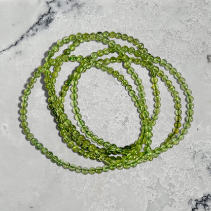 Four green Peridot crystal beaded bracelets arranged in overlapping loops on a white marble surface.