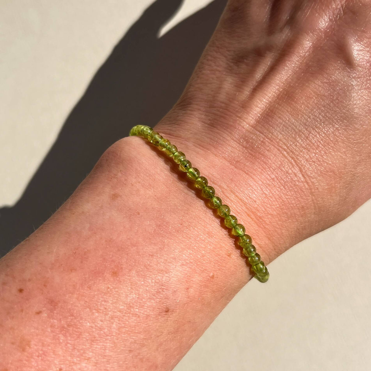 A close-up of a person's wrist wearing a thin round beaded bracelet made of translucent olive-green Peridot crystal beads, photographed in bright natural sunlight against a light beige background.