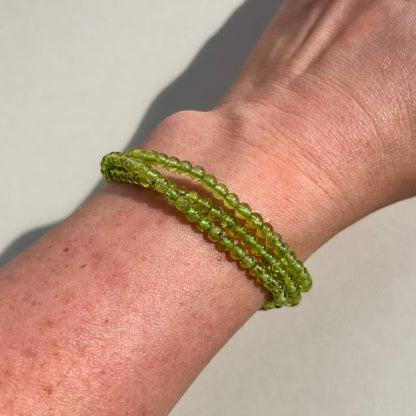 A close-up of a person's wrist wearing three Peridot crystal beaded bracelets in a translucent olive green colour, set against a neutral light gray background.