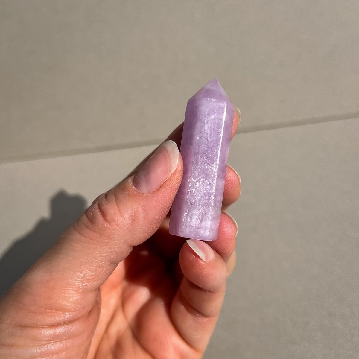 A hand holding a polished lilac pink shimmering kunzite crystal tower against a neutral gray tiled background.