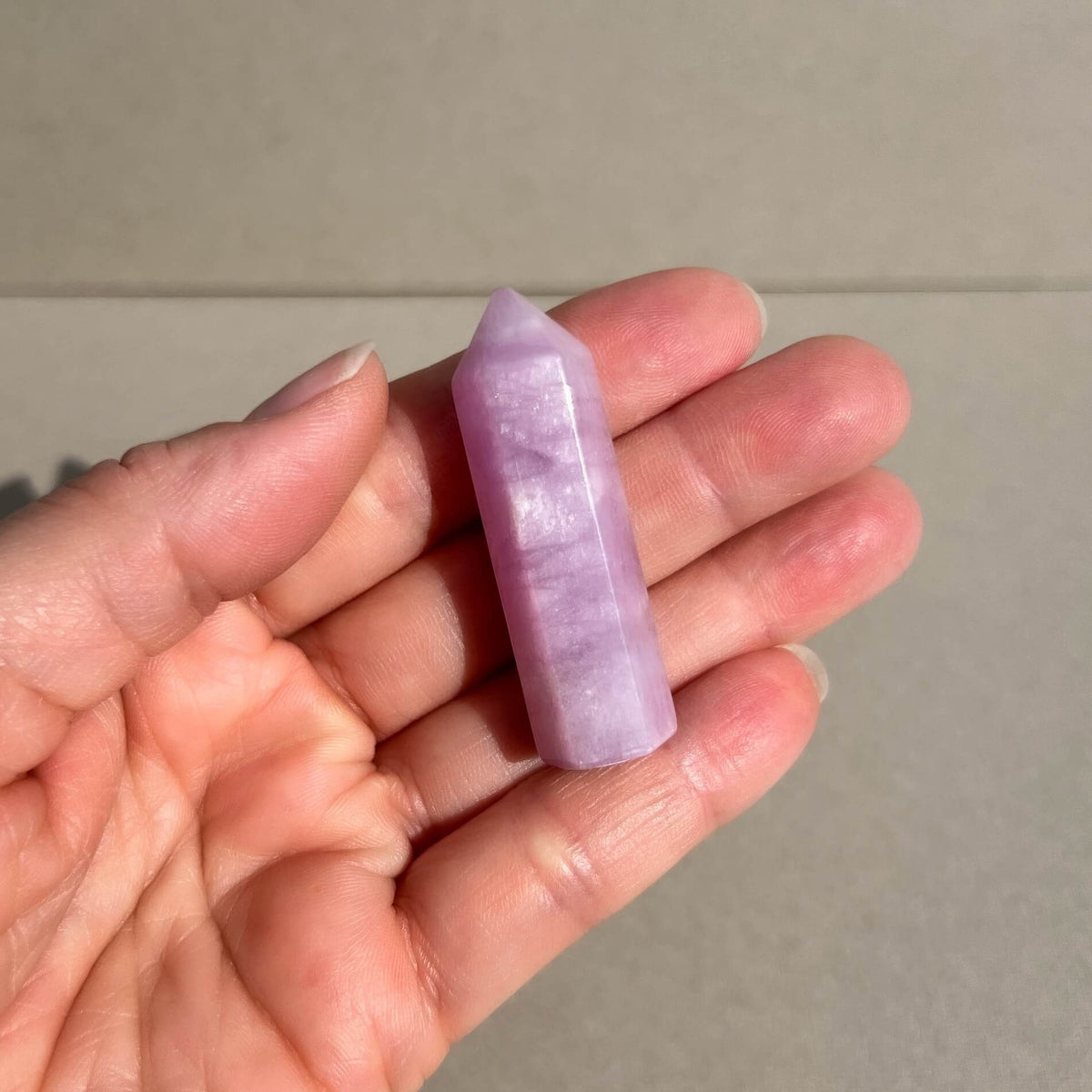 A small lavender-purple kunzite gemstone point held in a person's hand against a neutral gray background.
