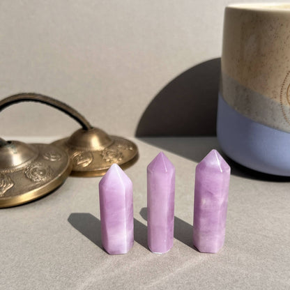 Three polished lilac pink kunzite crystal towers arranged upright on a neutral surface, with a ceramic candle and brass tingsha bells blurred in the background.