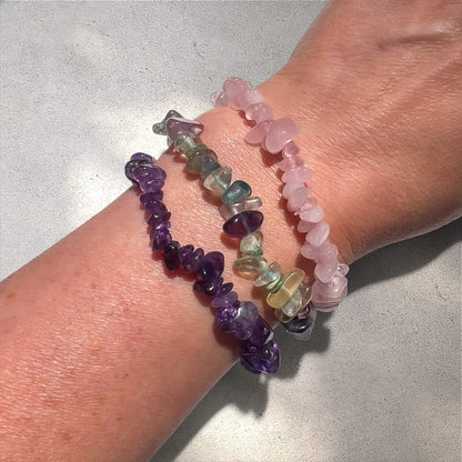 Stack of purple amethyst, rainbow fluorite and rose quartz crystal chip bracelets on a tanned woman's wrist against a natural sunlit background