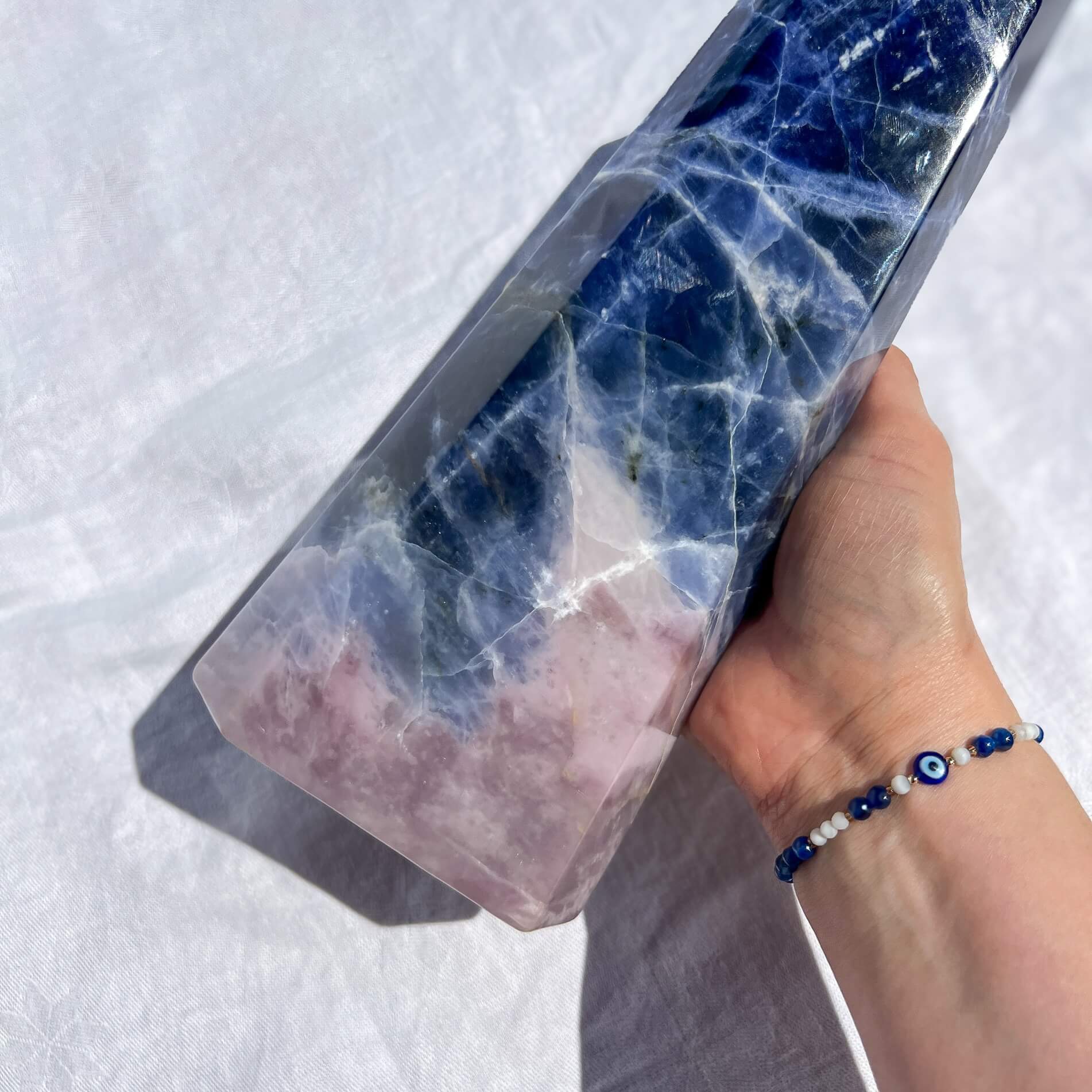 Hand wearing an evil eye bracelet holding a sodalite crystal tower with blue and pink marbled pattern against a white background