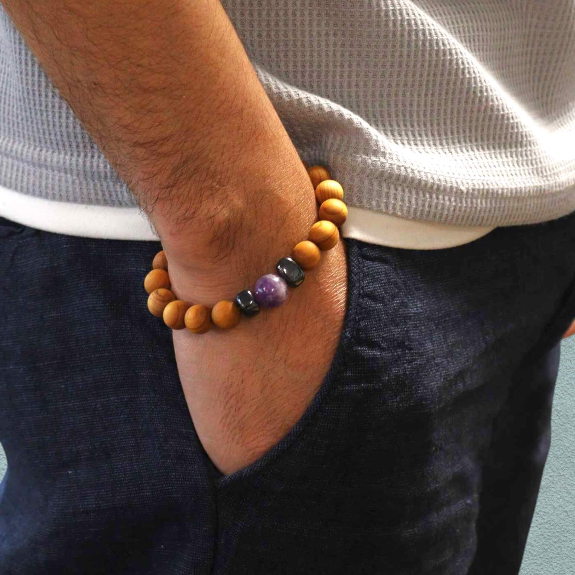 Man wearing a cedar wood bracelet with amethyst on an arm tucked in his jeans pocket, with a neutral background