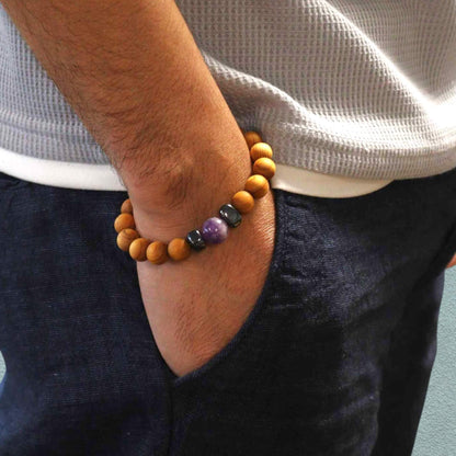 Man wearing a cedar wood bracelet with amethyst on an arm tucked in his jeans pocket, with a neutral background