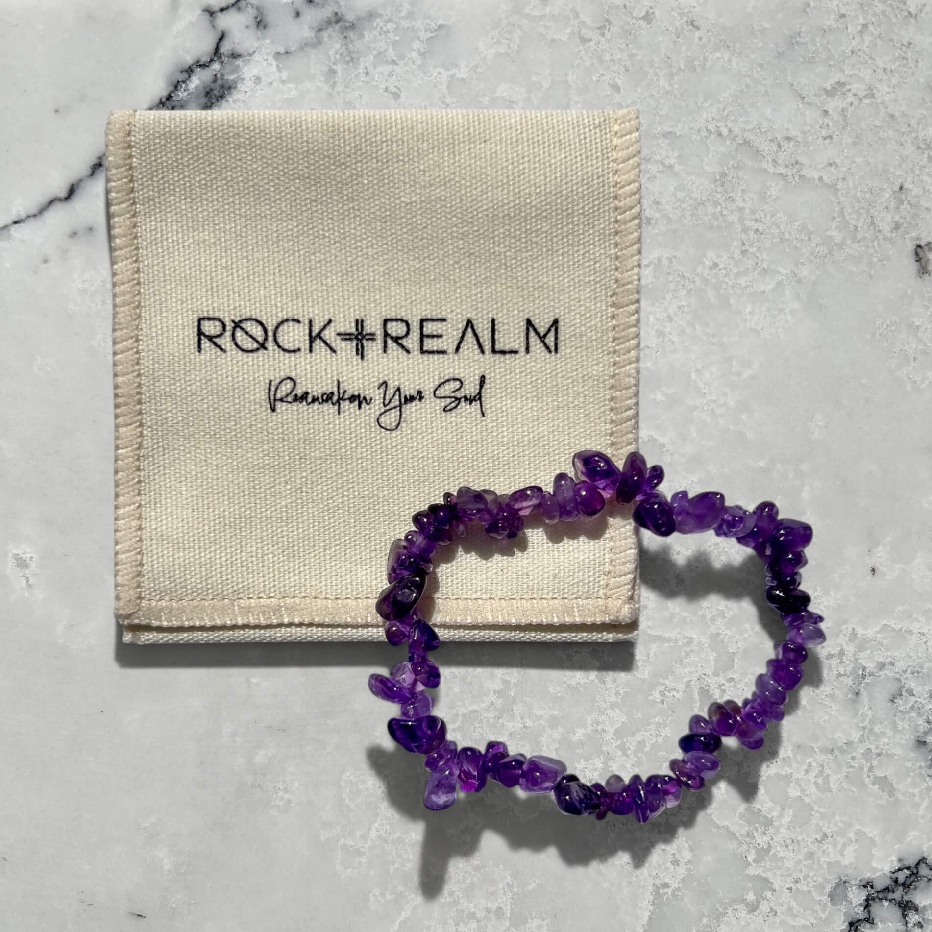 Purple amethyst crystal chip bracelet on a marble surface with 'Rock + Realm' branded cotton jewellery pouch