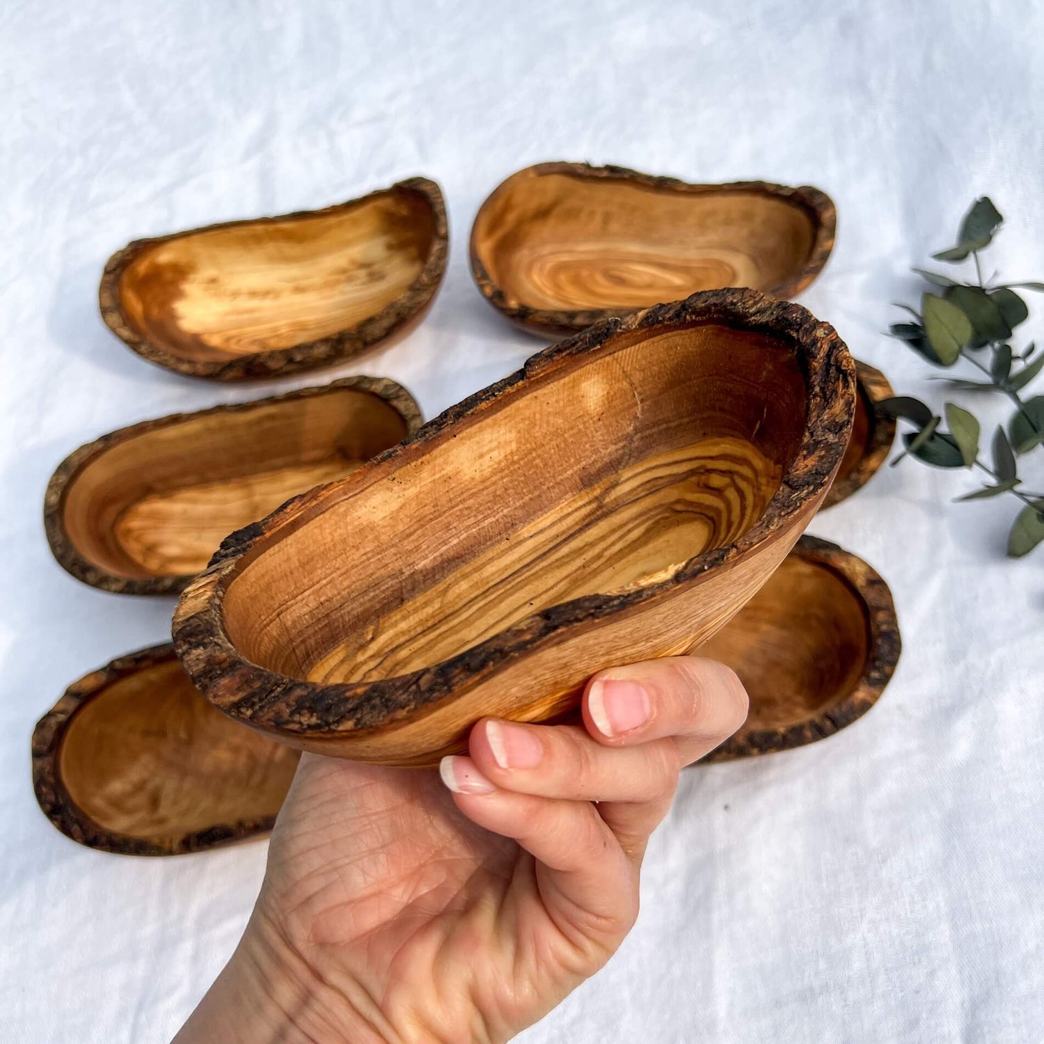 A raw edged hand carved rustic olive wood bowl is held in front of more hand carved wooden dishes