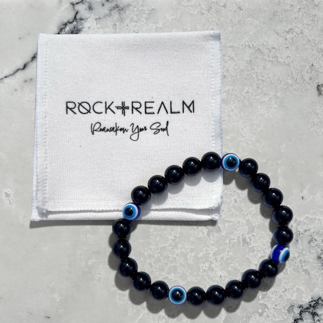 Black obsidian and blue evil eye bead bracelet on a white marble background with 'Rock + Realm' packaging.