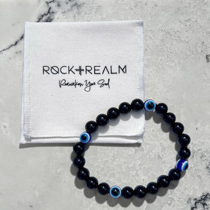 Black obsidian and blue evil eye bead bracelet on a white marble background with 'Rock + Realm' packaging.