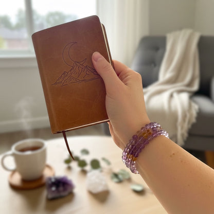 Rock + Realm Crystals ametrine beaded bracelet with purple and yellow stones, worn on a wrist holding a brown journal, a coffee table with amethyst, quartz and a steaming mug of tea in the background