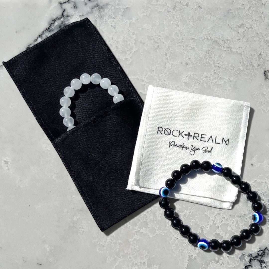 Rock + Realm branded white cotton pouch with a black obsidian beaded bracelet with blue evil eye charms and a white selenite beaded bracelet in a black cotton jewellery pouch.