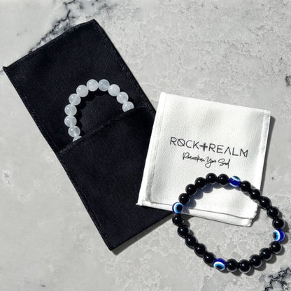 Rock + Realm branded white cotton pouch with a black obsidian beaded bracelet with blue evil eye charms and a white selenite beaded bracelet in a black cotton jewellery pouch.