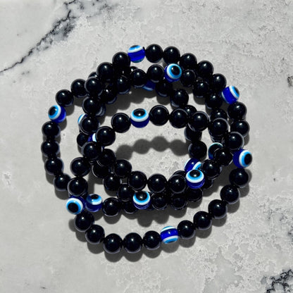 Stack of Rock + Realm Crystals black obsidian beaded bracelets with blue and white evil eye charms on a white marble surface