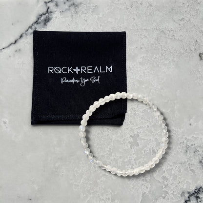 Rock + Realm Crystals delicate moonstone beaded stacking bracelet with tiny luminous beads and a black branded jewellery pouch on a marble surface
