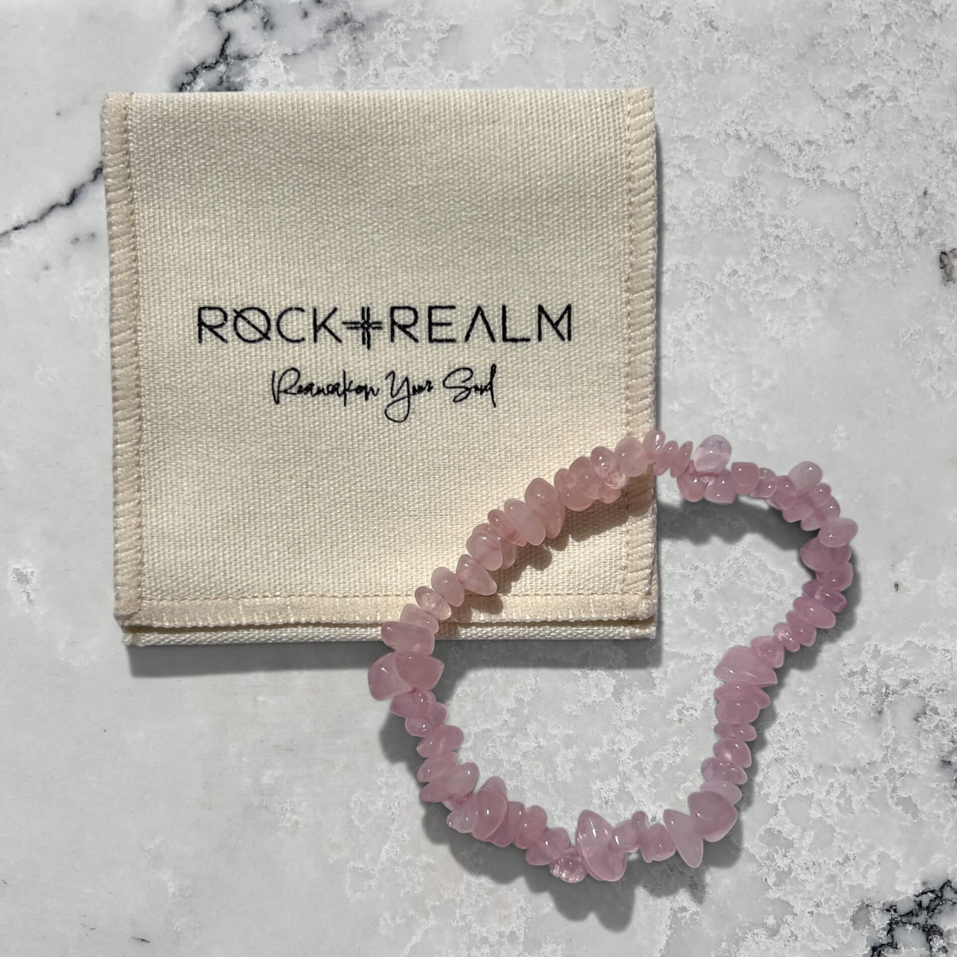 rose quartz crystal chip bracelet on a marble surface with 'Rock + Realm' pouch.