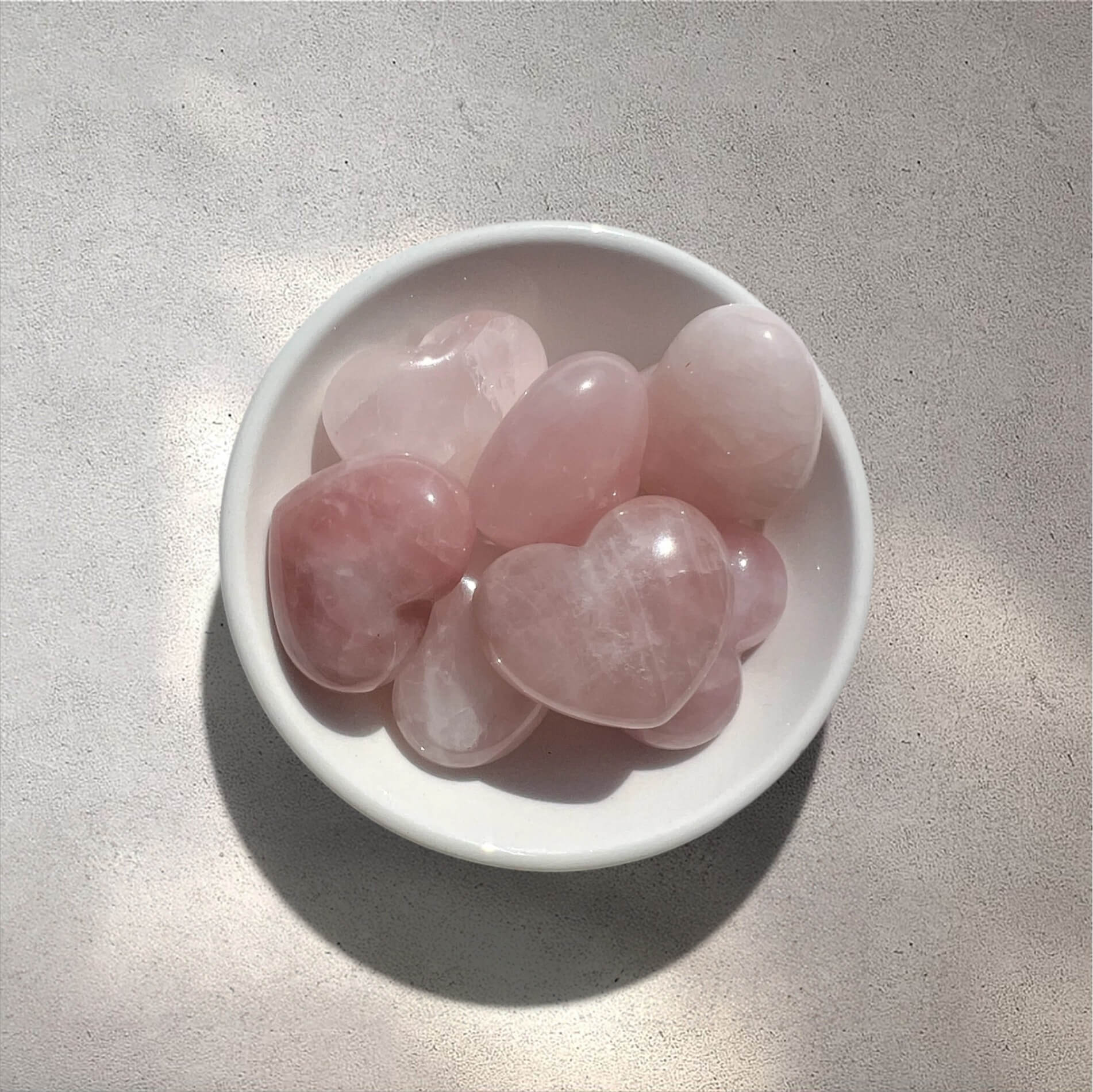 A small white ceramic dish filled with smooth, polished pink heart-shaped rose quartz crystals on a light grey textured surface.
