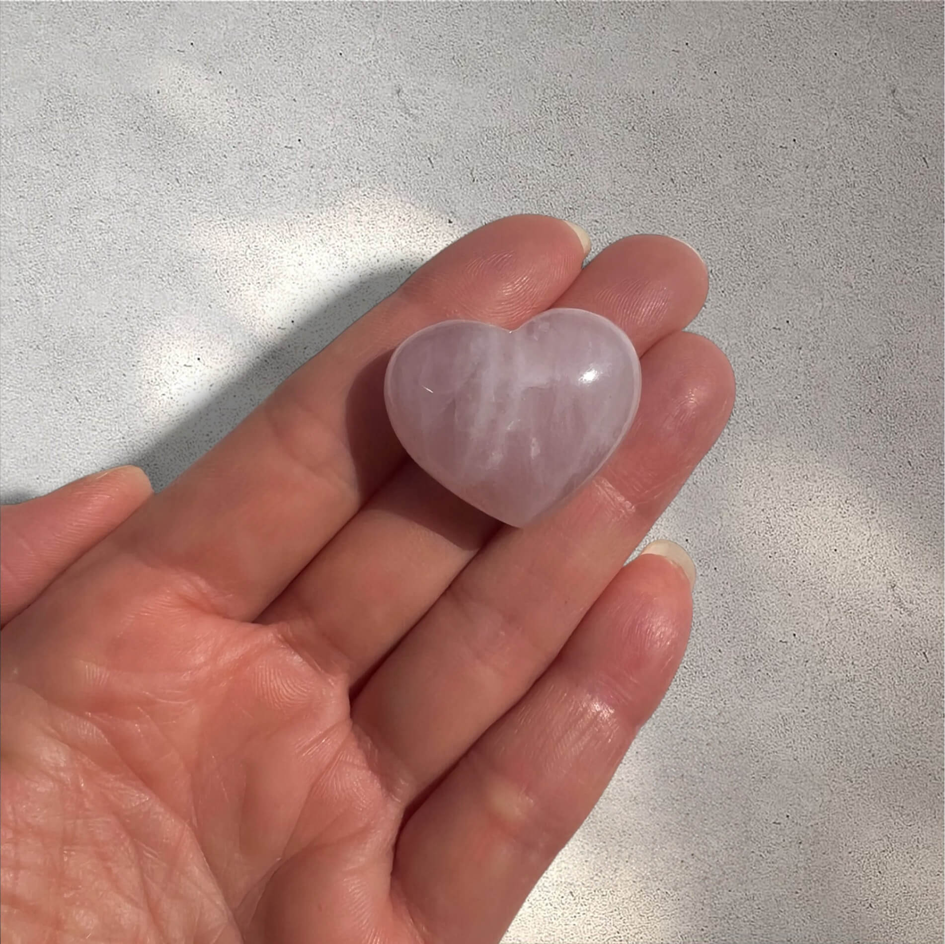 A small polished heart-shaped rose quartz crystal resting on a person's palm against a light grey textured background.