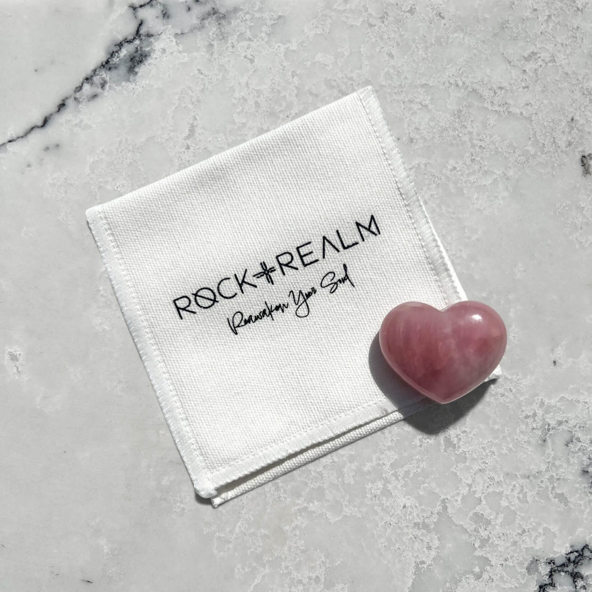 A white textured fabric pouch with black embroidered text reading 'ROCK + REALM Reawaken Your Soul' lies on a light marble surface next to a polished pink heart-shaped rose quartz crystal.