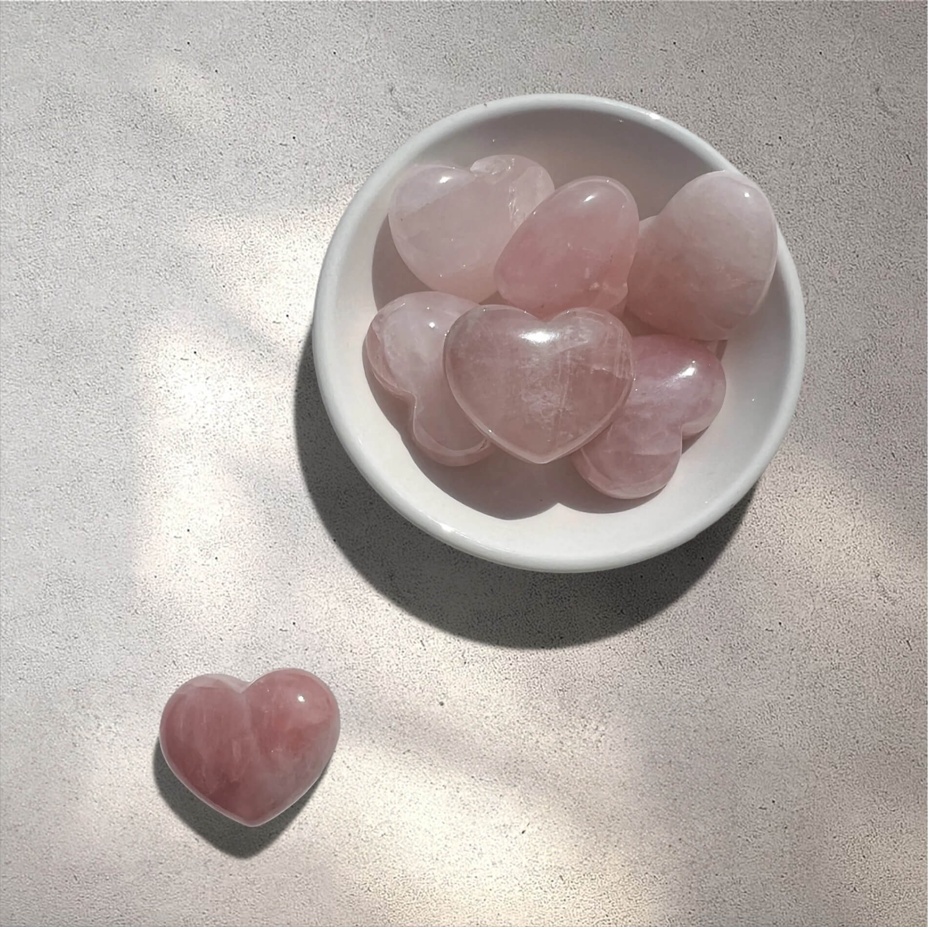Several polished pink heart-shaped rose quartz crystals are arranged in a small white ceramic dish, with one matching heart crystal placed beside it on a light textured surface.