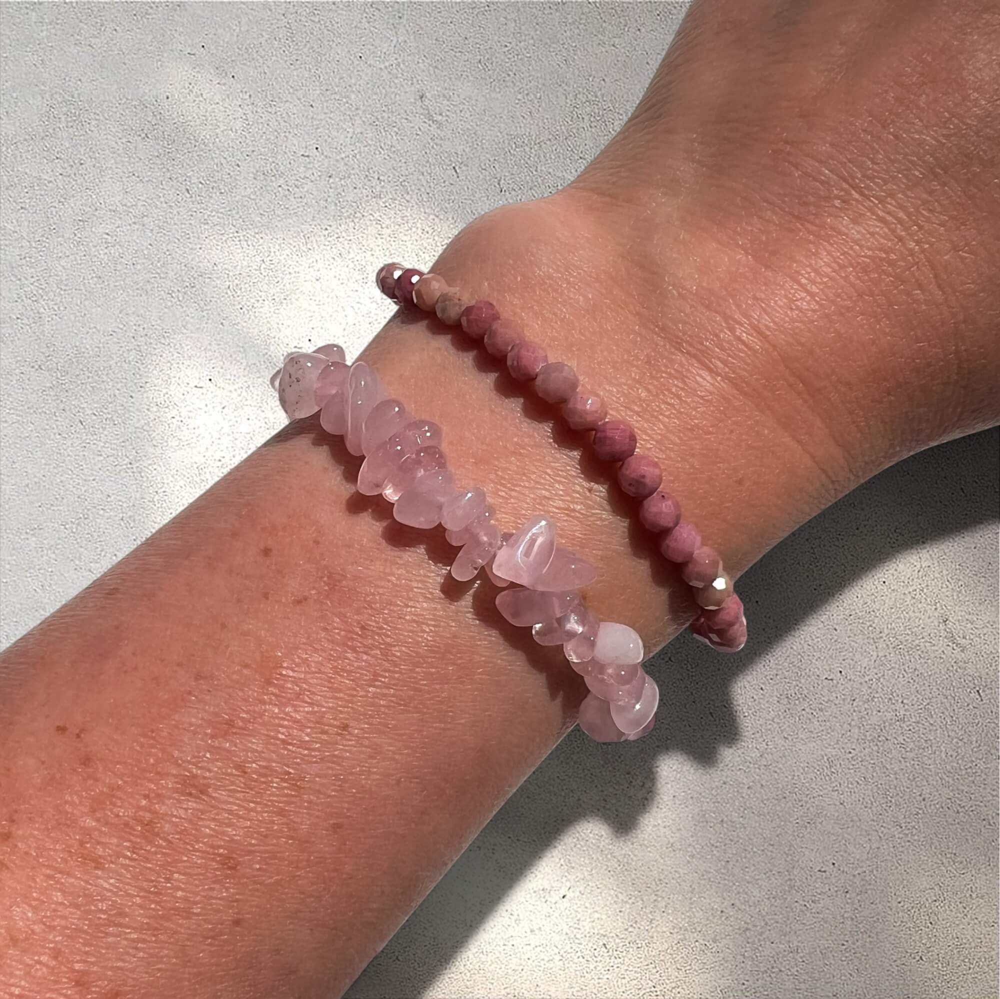 Close up of a wrist wearing a rose quartz crystal chip bracelet and a rhodonite faceted bead bracelet against a neutral background