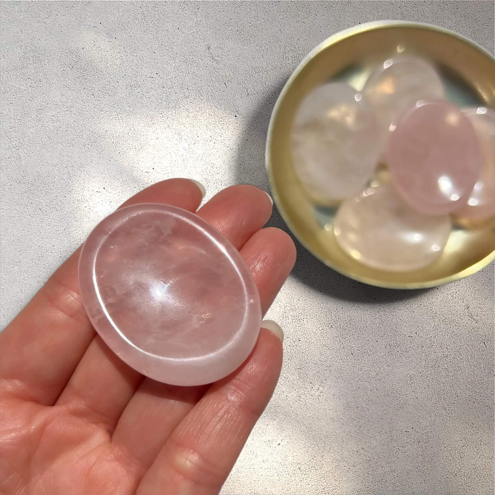 A hand holds a smooth, polished pink rose quartz worry stone, with a small gold dish containing several similar pink rose quartz thumb stones in the background.